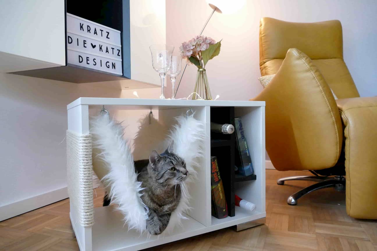 Katze liegt in der Hängematte eines breiten, weißen Kratzwürfels mit mehreren Regalfächern, in denen Bücher und Weinflaschen liegen und auf dem Blumen stehen.